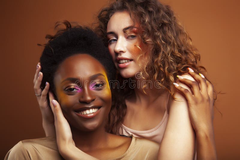 Two pretty girls african and caucasian blond posing cheerful together on brown background, ethnicity diverse lifestyle