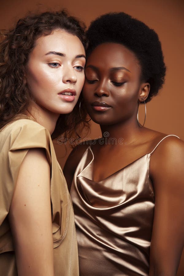 Two pretty girls african and caucasian blond posing cheerful together on brown background, ethnicity diverse lifestyle