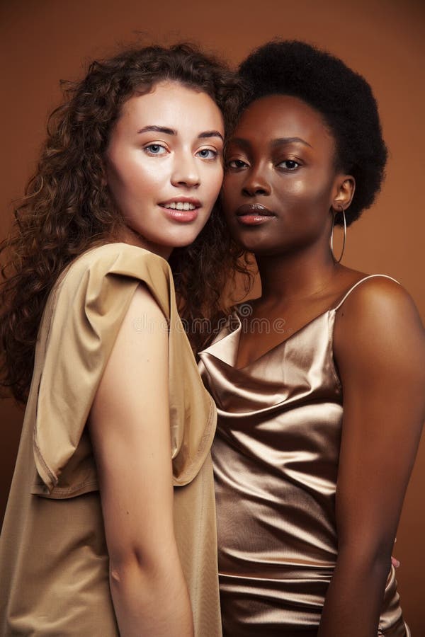 Two pretty girls african and caucasian blond posing cheerful together on brown background, ethnicity diverse lifestyle