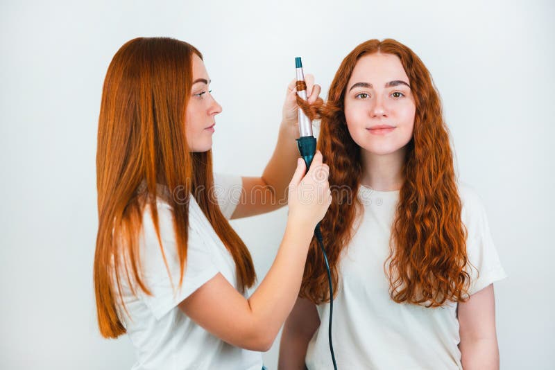 Two redheaded young women standing on isolated white backgroung, one curls her friend`s hair with styler, both look satisfied