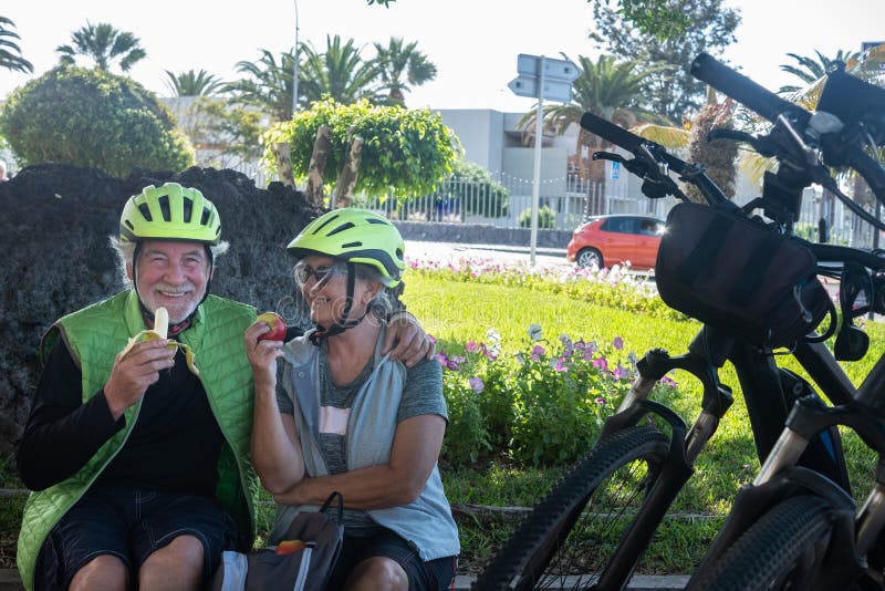 Two senior people cyclists with yellow helmet have a break in a green park eating a fruit and smiling. Moment of relaxation with