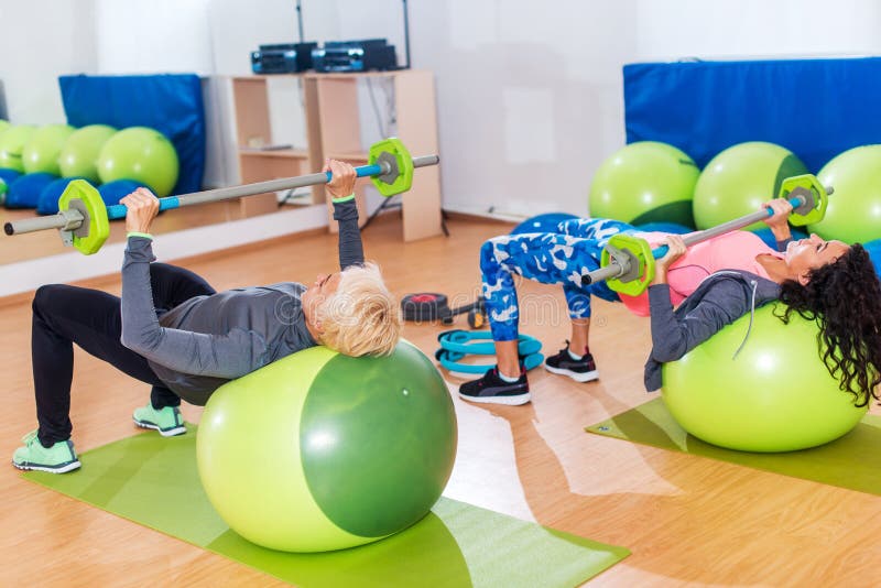 Two slim women exercising with barbell and Swiss ball in front of the mirror in training hall.