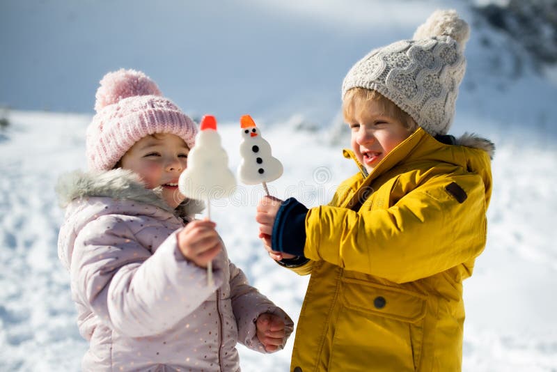 Two small children standing in snow in winter nature, eating sweets.