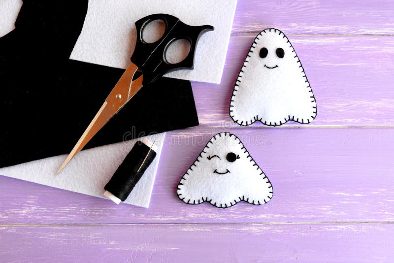 Two small white ghosts crafts, felt sheets, scissors, thread, needle on lilac wooden background. Hand Halloween decor idea