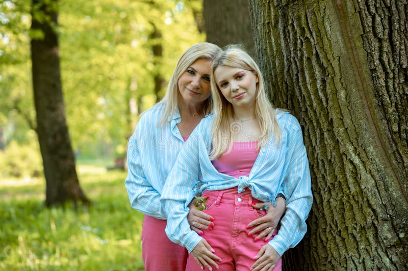 Two Smiling Blonde Mom And Daughter Dressed Identically Are Standing In Spring Park Near Tree