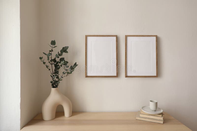 Two vertical picture frame mockup hanging on beige wall. Cup of coffee, books on wooden table. Vase with silver