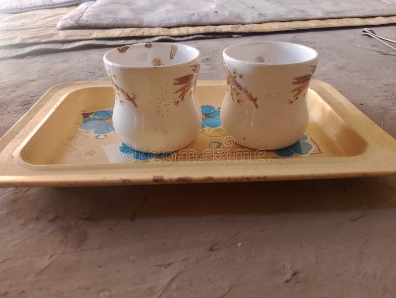 Two white ceramic cups with brown floral designs are placed on a rectangular yellow tray. The tray features a blue and white flower pattern in the center. The surface beneath appears to be a textured, dusty floor. In the background, there are faint outlines of fabric or coverings, suggesting a relaxed,. Ceram stock images, royalty-free photos and pictures