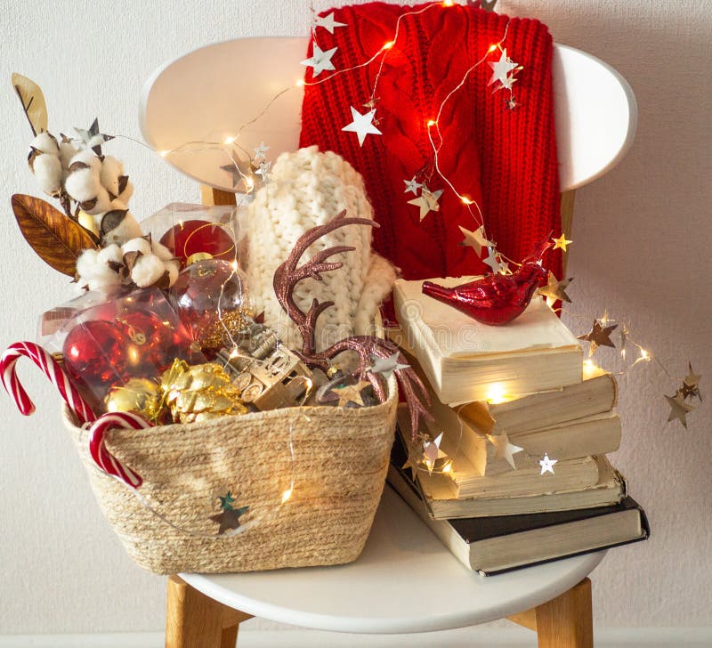 Two winter sweaters laid on a chair with a basket of Christmas decorations, books, led string lights. Winter reading. Winter mood