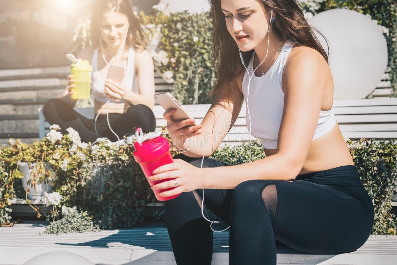 Two women athletes in sports clothes are sitting on bench, relax after sports training, use smartphones, listen to music