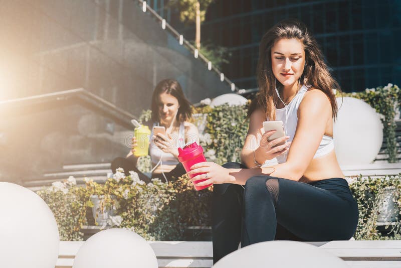 Two women athletes in sports clothes are sitting on bench, relax after sports training, use smartphones, listen to music stock images Two women athletes in sports clothes are sitting on bench, relax after sports training, use smartphones, listen to music stock images