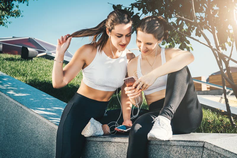 Two women athletes in sportswear sitting in park, relax after sports training, use smartphone, listening to music.