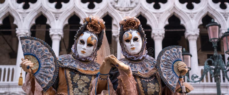 Two women in traditional costume and painted masks, with decorated fans, standing in front of the Doges Palace during Venice Carnival 2018