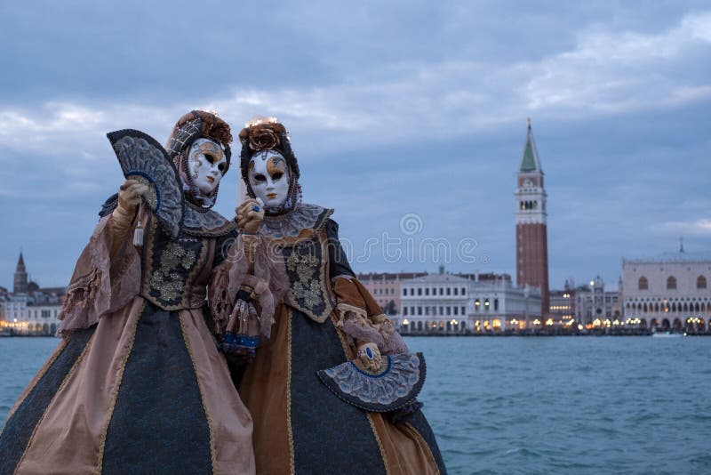 Two women in traditional costumes and masks, with decorated fans, at San Giorgio, with St Marks Square and bell tower behind