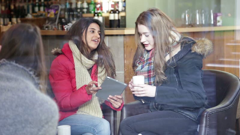 Two young women are sitting on a sofa in a cafe together. They are talking and using a digital tablet. Hot cafe video