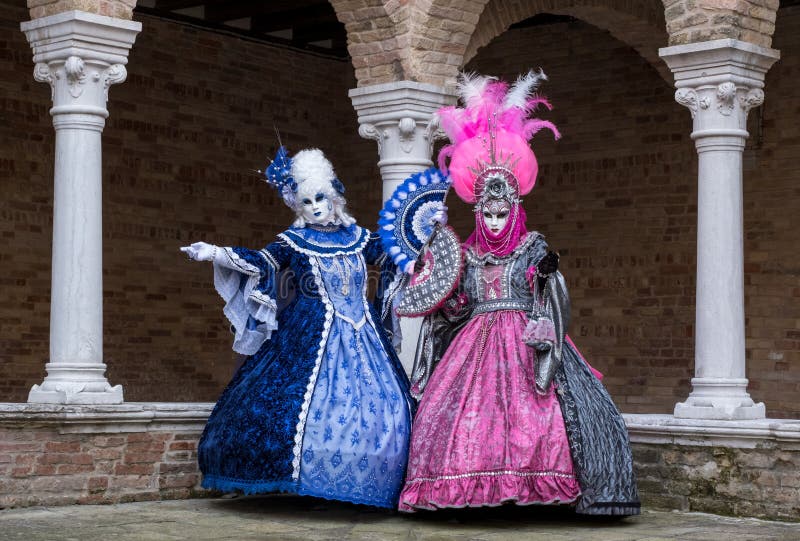 Two women wearing brightly colored masks and ornate blue and pink hand made costumes standing in the cloisters of a monastery during Venice Carnival Carnivale di Venezia, Italy. Two women wearing brightly colored masks and ornate blue and pink hand made costumes standing in the cloisters of a monastery during Venice Carnival Carnivale di Venezia, Italy.