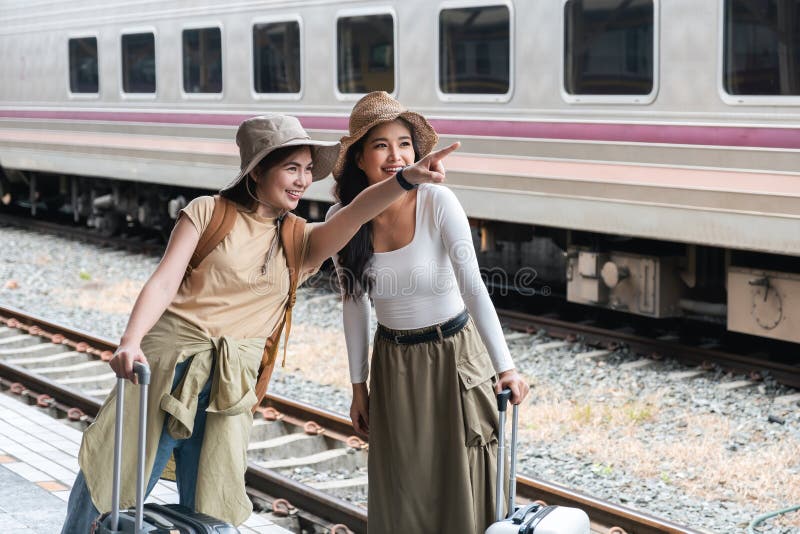Two young Asian women with backpacks and suitcases, wearing hats, pointing and smiling excitedly at a train station royalty free stock photography