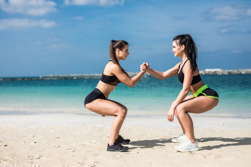 Strength in teamwork. Two young attractive women athletes exercise doing squats on the beach near beach stock photography Strength in teamwork. Two young attractive women athletes exercise doing squats on the beach near beach stock photography