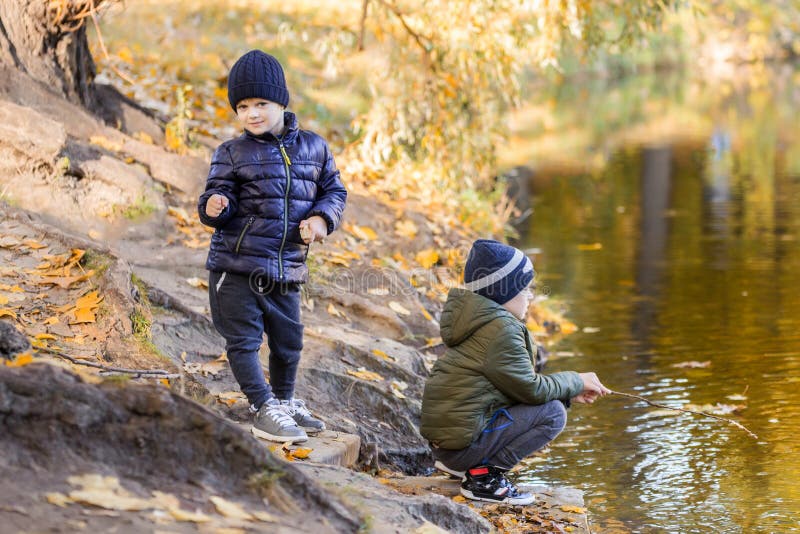 Two young boys playing fishing with sticks near pond in fall park. Little brothers having fun near lake or river in autumn. Happy