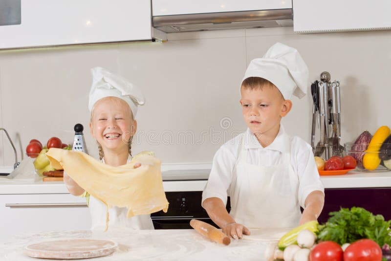 Two young children having fun making pizza with the little girl giggling as she holds aloft a large sheet of pastry that the little boy has just finished rolling out.