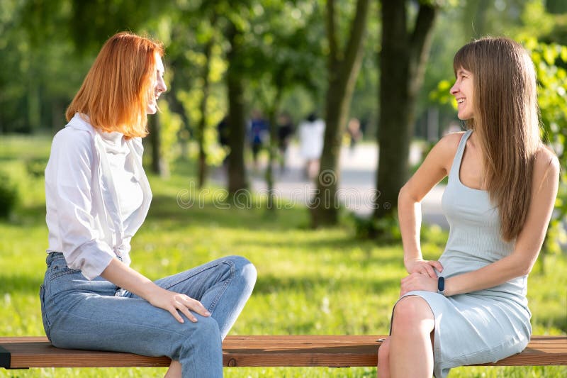 Two young girls friends sitting on a bench in summer park chatting happily having fun