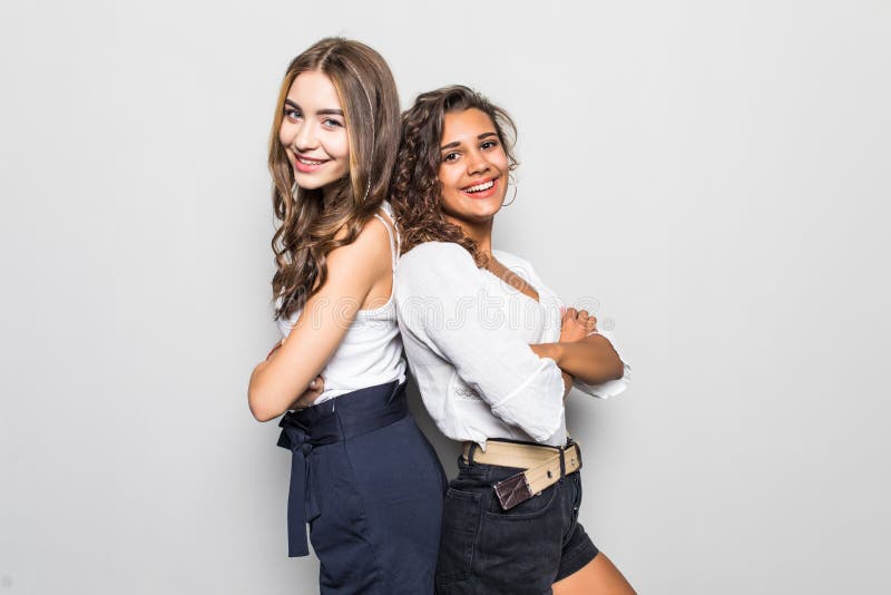 Two young girls stand back to back and smiling to camera on gray wall background