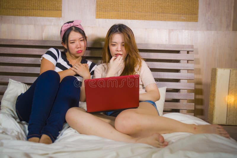 Two young pretty and happy Asian Chinese student girls together at home bedroom watching internet Korean drama with laptop