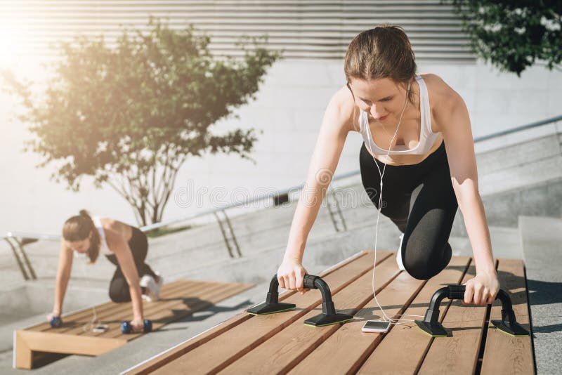 Two young smiling women, girls in sportswear doing exercises while listening to music. Workout, couching on city street. stock photos Two young smiling women, girls in sportswear doing exercises while listening to music. Workout, couching on city street. stock photos