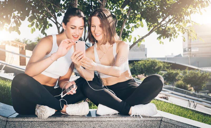 Two young women athletes in sportswear are sitting in park, relax after sports training, use smartphone. stock photography Two young women athletes in sportswear are sitting in park, relax after sports training, use smartphone. stock photography