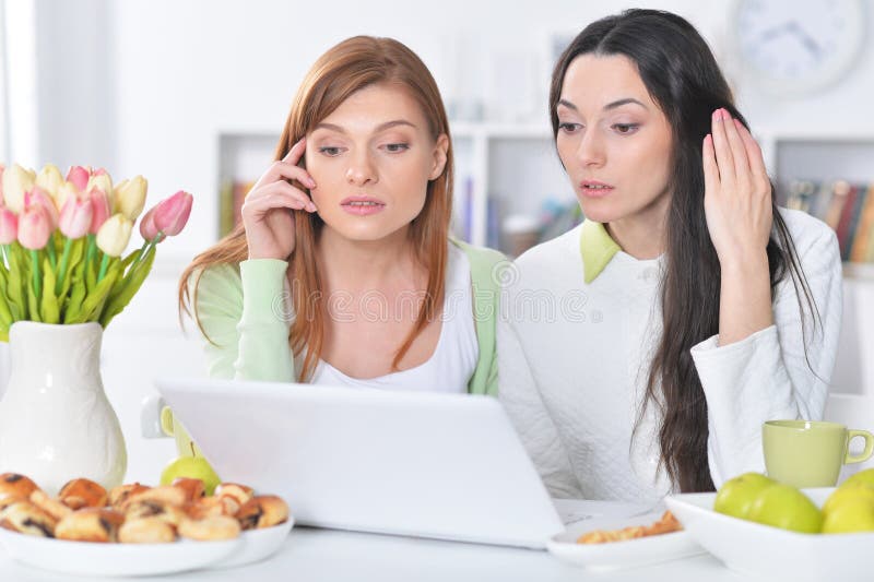 Portrait of two young women looking at laptop while sitting at kitchen table with cookies royalty free stock images Portrait of two young women looking at laptop while sitting at kitchen table with cookies royalty free stock images