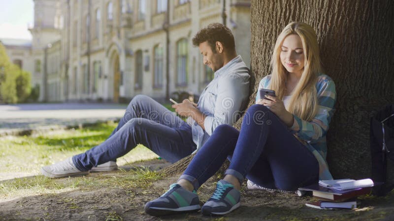 Type multiracial et fille s'asseyant sous l'arbre utilisant le sourire de téléphones portables, heureux photographie stock Type multiracial et fille s'asseyant sous l'arbre utilisant le sourire de téléphones portables, heureux photographie stock