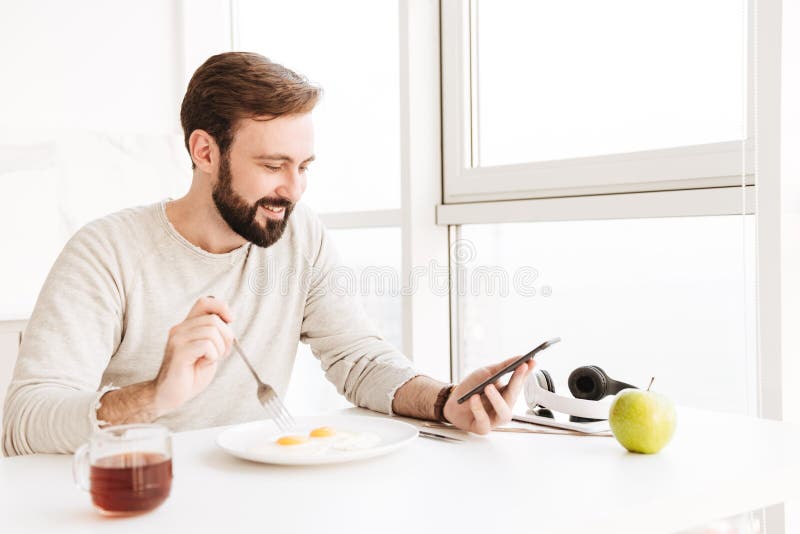 Type mûr heureux mangeant faisant frire des oeufs et à l'aide du téléphone portable, tandis que image libre de droits Type mûr heureux mangeant faisant frire des oeufs et à l'aide du téléphone portable, tandis que image libre de droits
