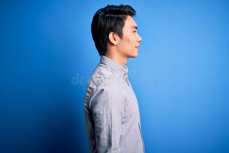 Tânăr chipeș chinez purtând cămașă obișnuită în picioare pe fond albastru izolat în căutarea unui profil relaxat lateral cu stock photography
