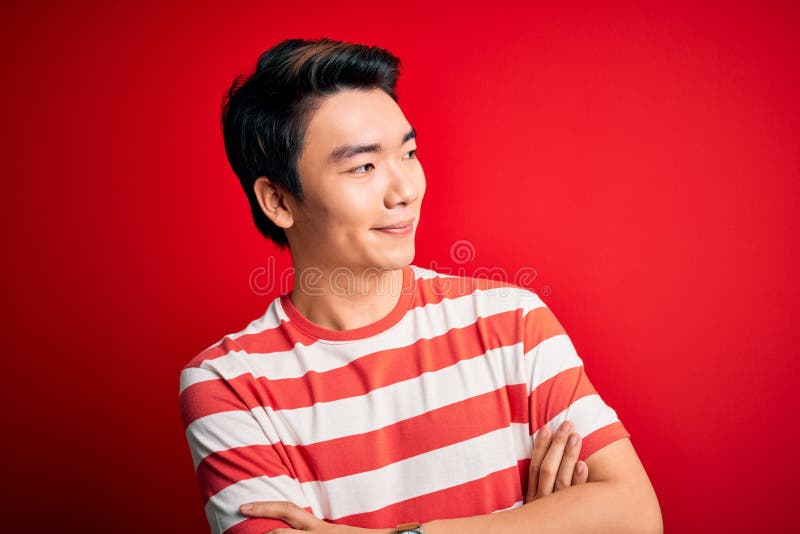 Tânăr chipeș chinez purtând o cămașă cu dungi, în picioare pe fond roșu, care se uită în lateral cu brațele încrucișate stock photography