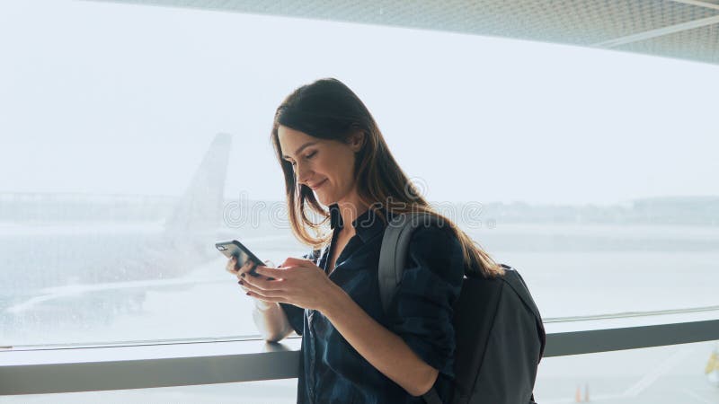 Tânără care utilizează smartphone lângă fereastra aeroportului. femeie europeană fericită cu rucsac utilizează aplicația mobilă în royalty free stock photo