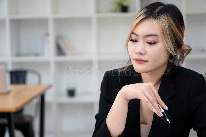 Tânără femeie de afaceri asiatică în negru, cu un pix care lucrează la un birou din birou stock images