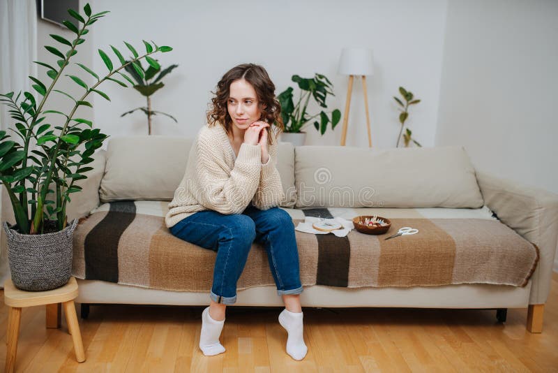Tänksam ung kvinna som sitter på soffan bredvid broderverktyg och material fotografering för bildbyråer