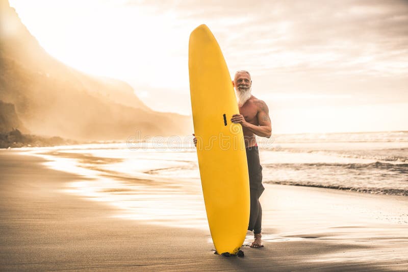 Tätowierter Senior Surfer, der Brandungsbrett auf dem Strand am glücklichen alten Mann des Sonnenuntergangs hat den Spaß macht, da lizenzfreie stockfotografie