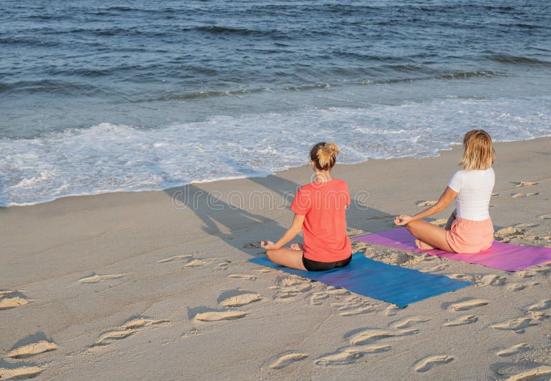 Título: Jovens mulheres que praticam a ioga na praia no por do sol Meditar das meninas, sentando-se na pose dos lótus imagem de stock Título: Jovens mulheres que praticam a ioga na praia no por do sol Meditar das meninas, sentando-se na pose dos lótus imagem de stock