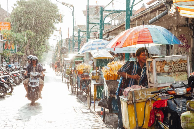 Ubud, Indonesia - March 08, 2016: Indonesian food vendor hides from the rain under umbrella of his stall on the street of Ubud, Bali, Indonesia on March 08, 2016. Ubud, Indonesia - March 08, 2016: Indonesian food vendor hides from the rain under umbrella of his stall on the street of Ubud, Bali, Indonesia on March 08, 2016