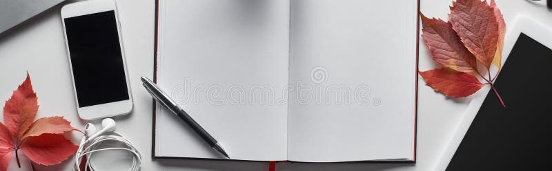Panoramic shot of blank notebook with pen near smartphone, digital tablet, earphones and red leaves of wild grapes on white table,stock image. Panoramic shot of blank notebook with pen near smartphone, digital tablet, earphones and red leaves of wild grapes on white table,stock image