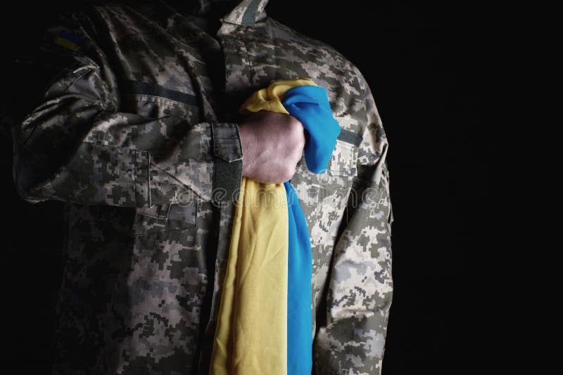 Ukrainian soldier holds in his hand the yellow-blue flag of the state, he pressed his hand to his chest. Concept of the memory of the dead soldiers stock photo Ukrainian soldier holds in his hand the yellow-blue flag of the state, he pressed his hand to his chest. Concept of the memory of the dead soldiers stock photo