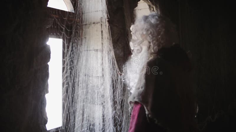 Um ancião com a barba branca longa no olhar vermelho da veste na janela fora do castelo