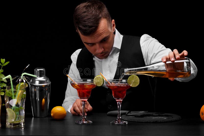 Um barman encantador derrama um cocktail nos vidros do margarita, fatias de cal, limão, fatias de cal em um fundo preto imagem de stock royalty free Um barman encantador derrama um cocktail nos vidros do margarita, fatias de cal, limão, fatias de cal em um fundo preto imagem de stock royalty free
