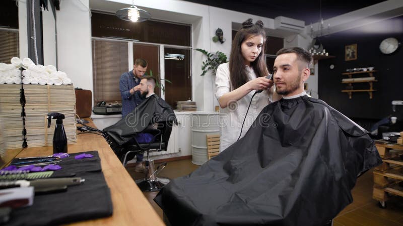 Um cabeleireiro da mulher nova e elegante está aparando o cabelo da parte mais inferior de sua cabeça para um salão de beleza