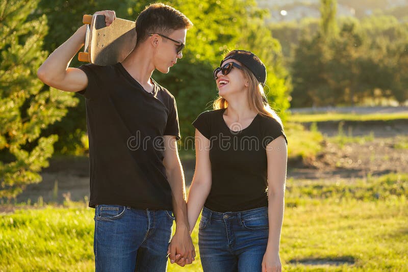 Um cara legal e uma garota adorável de óculos de sol, jeans e um skate de camiseta preta segurando as mãos no parque de verão fotos de stock royalty free Um cara legal e uma garota adorável de óculos de sol, jeans e um skate de camiseta preta segurando as mãos no parque de verão fotos de stock royalty free