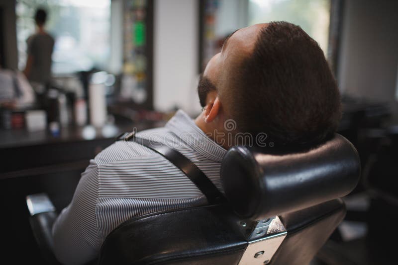 Um close-up de um homem considerável que senta-se em uma cadeira do ` s do cabeleireiro Um homem à moda novo com uma barba em um imagem de stock Um close-up de um homem considerável que senta-se em uma cadeira do ` s do cabeleireiro Um homem à moda novo com uma barba em um imagem de stock
