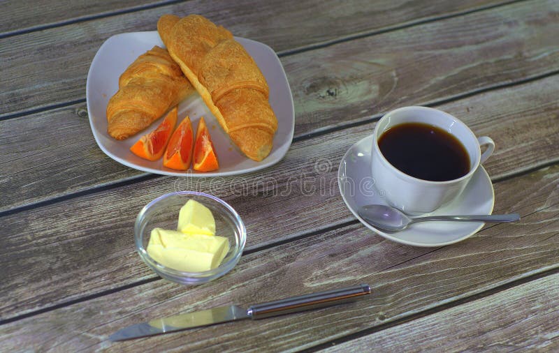 Um copo do café preto em uns pires com uma colher, uma placa com croissant e fatias do limão com suporte da manteiga em uma tabel fotografia de stock Um copo do café preto em uns pires com uma colher, uma placa com croissant e fatias do limão com suporte da manteiga em uma tabel fotografia de stock