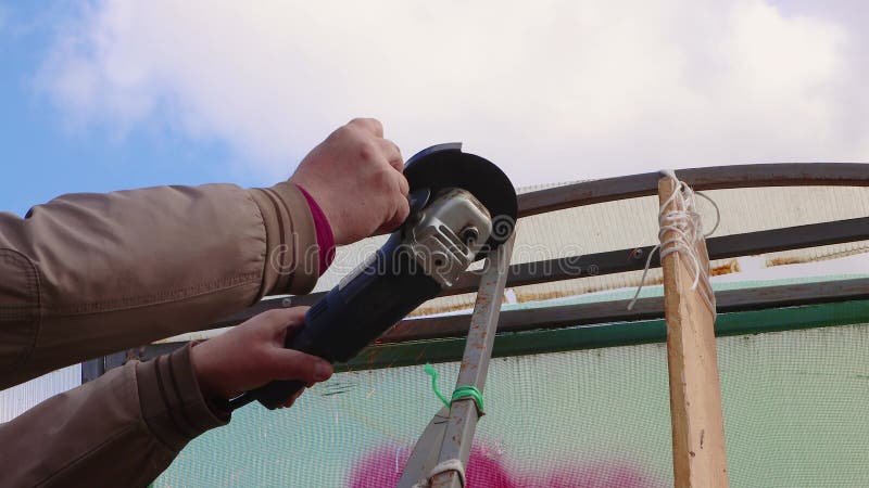 Um homem com uma afiadora angular corta uma estufa de metal enferrujada contra um céu azul.