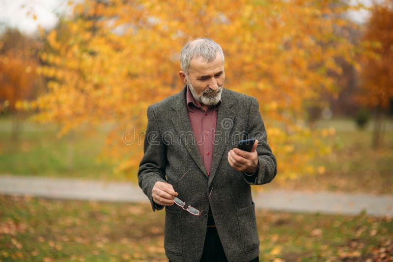 Um homem idoso considerável com a barba nos vidros está usando um telefone Caminhada no parque no outono fotos de stock Um homem idoso considerável com a barba nos vidros está usando um telefone Caminhada no parque no outono fotos de stock
