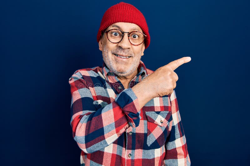 Handsome mature man wearing hipster look with wool cap cheerful with a smile of face pointing with hand and finger up to the side with happy and natural expression on face. Handsome mature man wearing hipster look with wool cap cheerful with a smile of face pointing with hand and finger up to the side with happy and natural expression on face